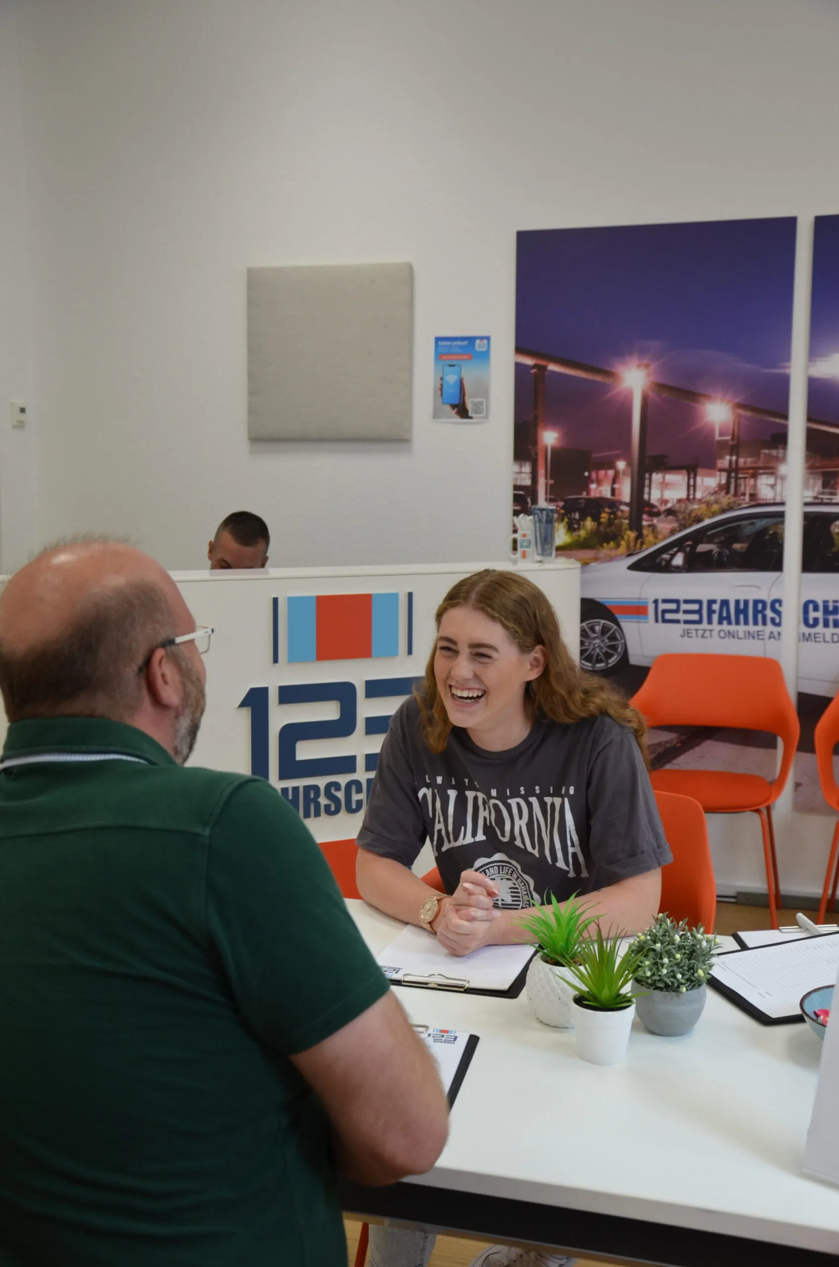 Fahrlehrer der 123Fahrschule sitzt mit Fahrschülerin an einem Tisch. Im Hintegrund ist die Rezeption der Fahrschule zu sehen